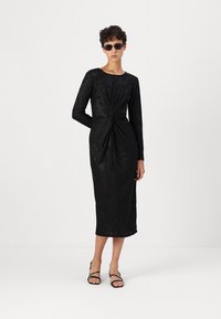 Vestido preto de manga longa com padrão floral texturizado, detalhe frontal torcido, silhueta ajustada, até aos joelhos, combinado com sandálias pretas de tiras e óculos de sol.