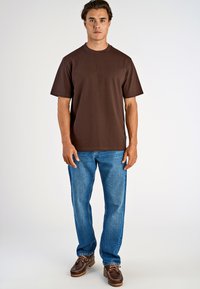 Brun kortärmad t-shirt med rund hals, ihop med blå jeans och bruna båt-skor, stående mot en plain ljus bakgrund.