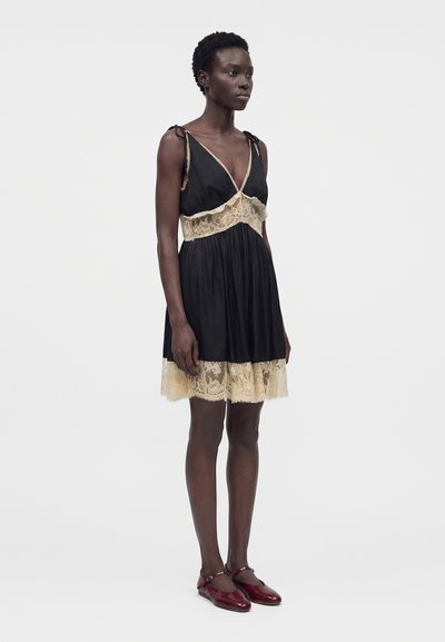 Γυναίκα που στέκεται, φοράει μαύρο αμάνικο φόρεμα με μπεζ δαντέλα και κόκκινα παπούτσια Mary Jane, με λευκό φόντο.