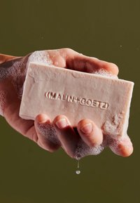 Ein rechteckiges, blassrosa Stück Seife mit geprägt eingestanztem Logo, gehalten in einer mit Blasen bedeckten Hand, aus der ein Wassertropfen fällt.