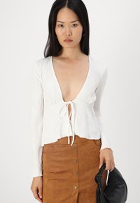 Haut long à manches blanches côtelé avec un décolleté en V profond et un détail de nœud à l'avant, associé à une jupe en daim brun avec des accents de boutons et un sac noir.