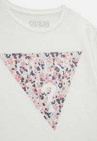 T-shirt en coton blanc avec un grand triangle à motifs floraux portant le mot "GUESS" en rose clair et un point d'interrogation au centre.