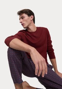Red long-sleeve shirt paired with patterned pants in dark burgundy, featuring blue and white geometric shapes. Model seated with one hand resting.
