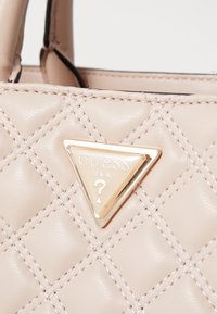 Borsa a tracolla in pelle beige trapuntata con una placca triangolare in metallo oro con la scritta "GUESS U.S.A." e un dettaglio a forma di punto interrogativo.