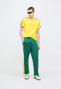 Gelbes T-Shirt mit großem, strukturiertem adidas-Logo, kombiniert mit grünen, gemusterten Trainingshosen mit gelben Seitenstreifen. Schwarze Sonnenbrille.