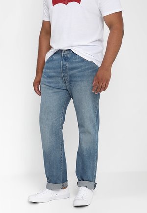 Hombre con vaqueros azul claro remangados, camiseta blanca con gráfico y zapatillas blancas, de pie frente a un fondo liso.