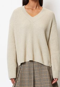 Maglione a coste beige con scollo a V e spalle abbattute, caratterizzato da una vestibilità rilassata e un orlo corto. Abbinato a una gonna a quadretti.
