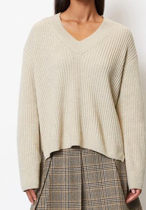 Maglione a coste beige con scollo a V e spalle abbattute, caratterizzato da una vestibilità rilassata e un orlo corto. Abbinato a una gonna a quadretti.