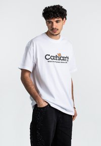 Camiseta de algodón blanca con el logo y texto en negro "Carhartt", corte relajado y mangas cortas. Combinada con pantalones negros con detalles bordados.