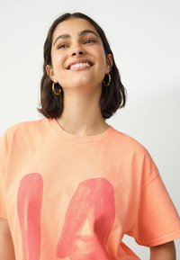 Overdimensioneret koral T-shirt med et stort pink grafisk design på forsiden. Korte ærmer, bomuldsstof og rund halsudskæring.