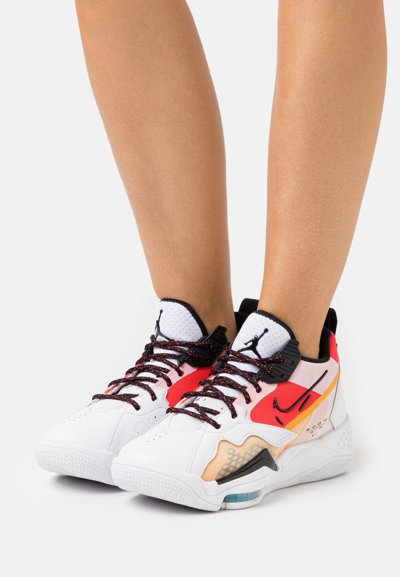 Bílé sportovní boty s červenými, černými a oranžovými akcenty; texturovaný svršek, síťovaná špička, černé tkaničky a viditelné detaily vzduchového polstrování.