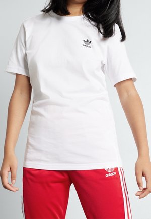 Person iført en hvid Adidas t-shirt og røde Adidas træningsbukser med hvide striber, stående mod en lys baggrund.