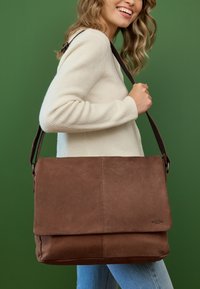 Bolso mensajero de cuero marrón con solapa plegable, correa de hombro ajustable y detalles de costura visibles. Llevado por un modelo frente a un fondo verde.