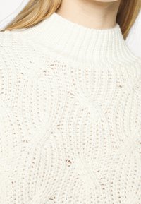 Chunky knit sweater in off-white, featuring a high ribbed collar and intricate wave patterns across the textured fabric.