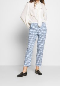 Blouse blanche avec un détail nœud papillon, associée à un pantalon bleu clair à motifs géométriques. Des mocassins noirs complètent la tenue.