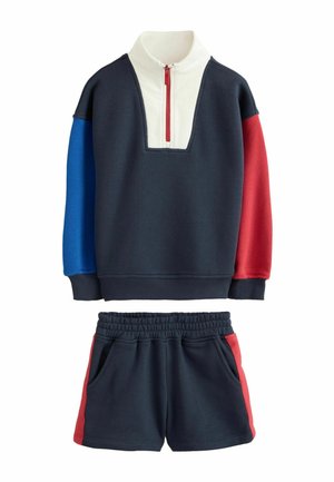 Marineblaue Sweatshirt mit blau-roten langen Ärmeln, weißem Kragen mit rotem Reißverschluss, kombiniert mit marineblauen Shorts mit roten Seitenstreifen.