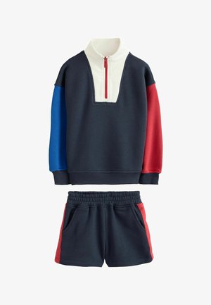 Marineblaue Sweatshirt mit blau-roten langen Ärmeln, weißem Kragen mit rotem Reißverschluss, kombiniert mit marineblauen Shorts mit roten Seitenstreifen.