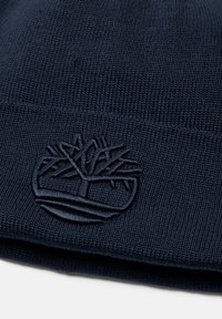 Marinblå stickad mössa med texturerat mönster och påsytt logotypdesign som föreställer ett träd. Tillverkad av mjukt, elastiskt material för komfort.