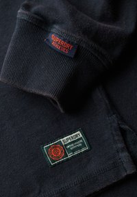 Zwarte katoenen sweatshirt met ribgebreide manchetten, twee labels: "Superdry Athletics" in oranje en "Superdry Vintage Athletic Outfitters" in groen.