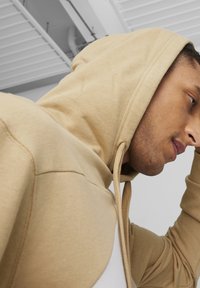 Beżowa bluza z kapturem i sznurkiem, wyró�żniająca się miękką fakturą i luźnym krojem. Widok z boku pod kątem, ukazujący materiał i subtelne detale przeszycia.