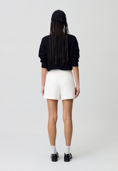 Suéter negro, pantalones cortos blancos, gorra negra, cabello largo trenzado, zapatos negros y calcetines grises. Diseño minimalista con colores neutros.