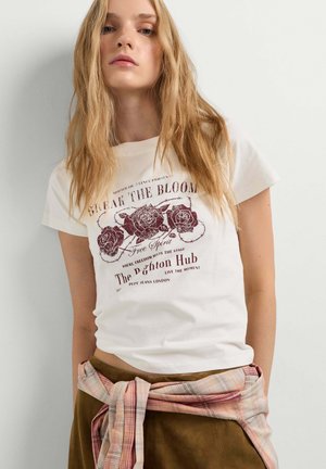 Joven mujer con cabello largo y rubio, que lleva una camiseta gráfica blanca con diseño de flores y texto, pantalones marrones y una camisa de cuadros atada alrededor de su cintura.