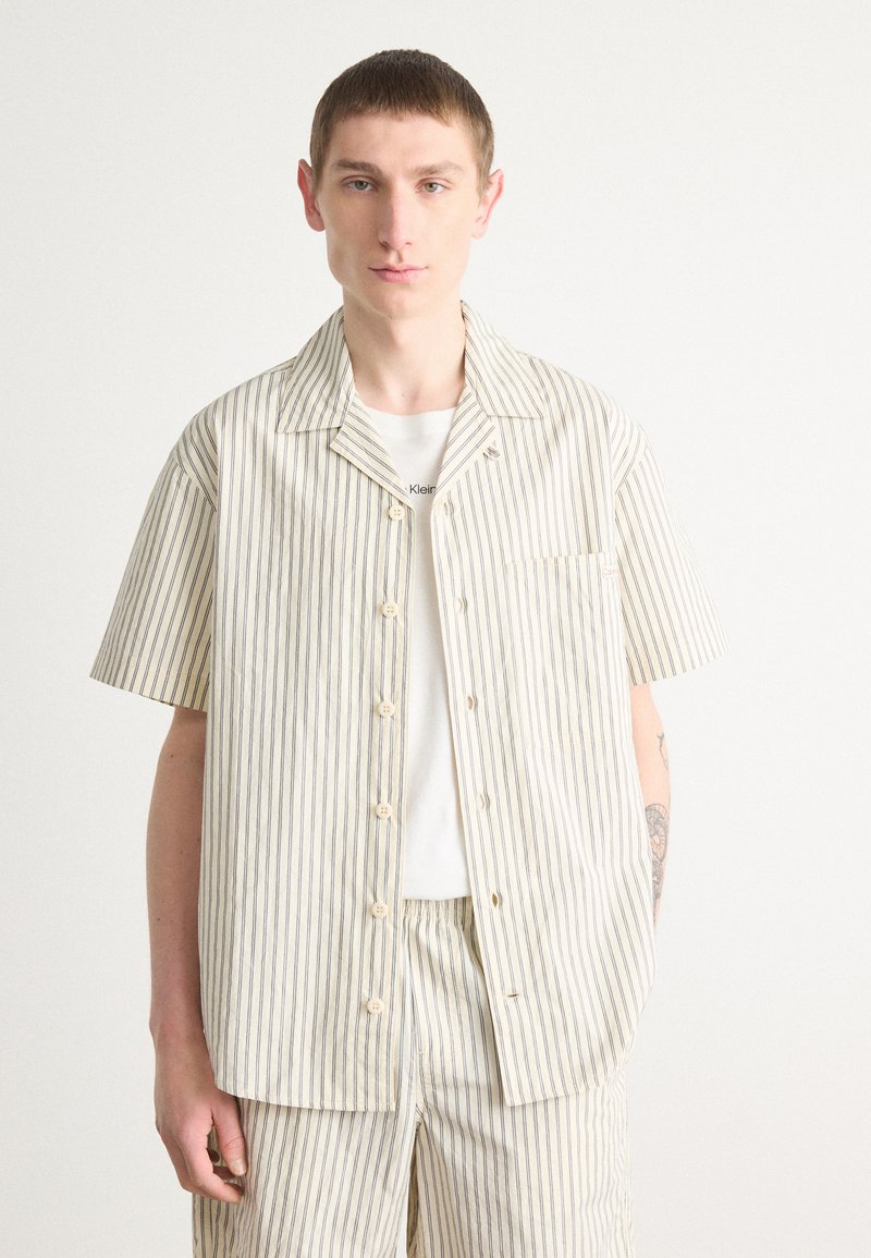 Korte mouwen, knoopshirt in crème met marineblauwe verticale strepen. Heeft een borstzak en is gemaakt van lichte stof.