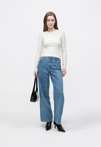 Top bianco a maniche lunghe con motivo a rilievo, abbinato a jeans blu a vita alta e gamba larga. La modella tiene una borsa nera e indossa tacchi neri.