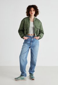 Zkrátená zelená bomber bunda s dvoma prednými vreckami, šedý zkrátený top, svetlo modré džínsy a mätovo zelené tenisky s hnedými akcentmi.