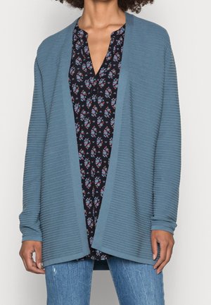 Woman wearing a ribbed blue open-front cardigan over a black floral blouse with blue and red accents, paired with blue jeans.