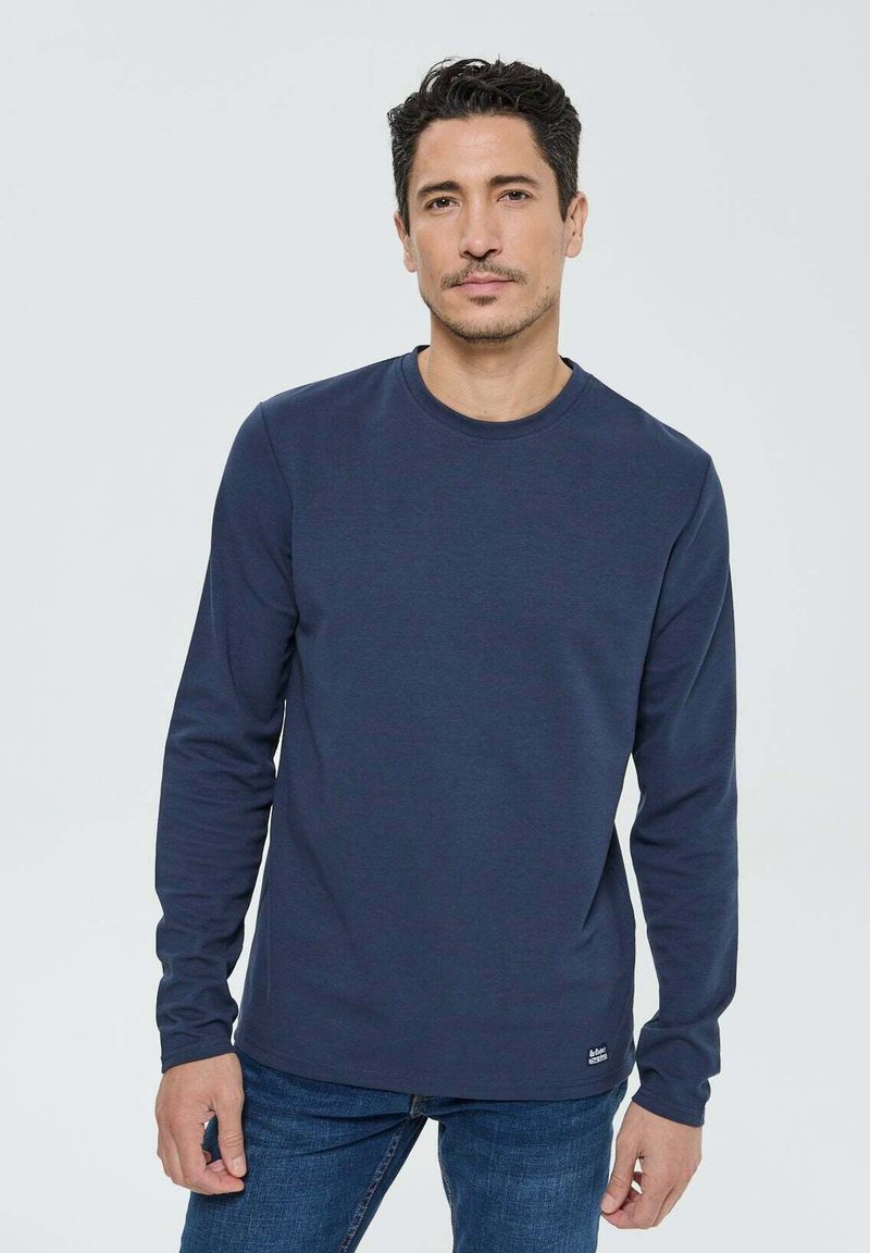 Homme aux cheveux courts et foncés portant une chemise à manches longues bleu marine et un jean bleu, se tenant devant un fond gris clair uni.