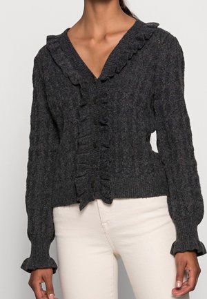 Nainen pukeutunut tummanharmaaseen palmikkoneuleiseen neulejakkuun, jossa on röyhelöinen V-pääntie, napillinen suljenta ja röyhelöiset hihansuut, yhdistettynä vaaleanbeigeihin housuihin.