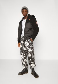 Schwarze gepolsterte Jacke mit Fleece-Akzenten, weißes T-Shirt, schwarz-weiße gemusterte Hose mit Gesichtern und schwarze Stiefel mit gelben Nähten.