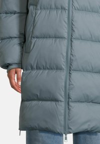 Nahaufnahme einer Person, die eine graue Daunenjacke mit einem Reißverschluss vorne und Seitentaschen trägt, während unten blaue Jeans sichtbar sind.