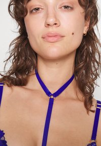 Arnês roxo com correias e um acréscimo circular dourado. O material é liso com um acabamento ligeiramente brilhante. Detalhes intrincados nas laterais.