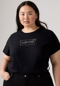 T-shirt en coton noir avec un logo Levi's brodé blanc sur la poitrine, associé à un jean noir taille haute. Le mannequin a de longs cheveux foncés.