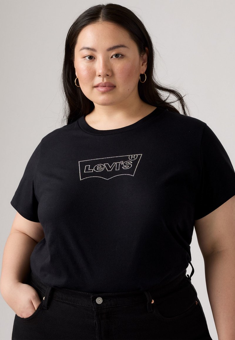 T-shirt en coton noir avec un logo Levi's brodé blanc sur la poitrine, associé à un jean noir taille haute. Le mannequin a de longs cheveux foncés.