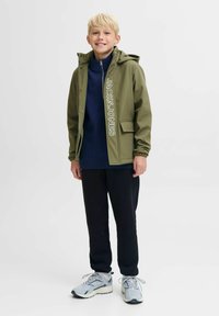 Veste vert olive avec capuche et lettrage blanc, superposée sur un pull bleu marine, associée à un pantalon noir et des baskets gris clair.
