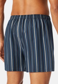 Hombre con pantalones cortos elásticos de cintura alta a rayas verticales en azul y gris, visto desde atrás, con torso y brazos desnudos visibles.