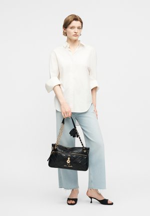 Γυναίκα που φοράει λευκό πουκάμισο με κουμπιά και ανοιχτό μπλε ξεθωριασμένο τζιν κρατώντας μια μαύρη καπιτονέ τσάντα με χρυσές αλυσίδες.