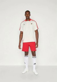 Sudadera blanca a rayas con acentos rojos, pantalones cortos rojos y calcetas blancas con rayas negras. El modelo está de pie sobre un fondo neutro.