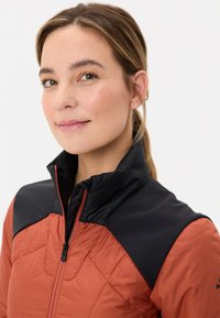 Gequilted, roest-oranje jas met zwarte kraag en schouders. Voorzien van een hoge nek en ritsluiting. Glad textuur met contrasterende patronen.