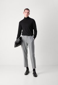 Svart turtleneck, grå plisserte bukser og svarte sko. Modellen holder en svart jakke. Glatt stoff, skreddersydd passform, nøytrale farger.