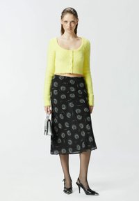 Žuti krzneni skraćeni kardigan s dugim rukavima, uparen s crnom midi suknjom s uzorkom i zavojitim detaljima, crnim štiklama i malom srebrnom torbicom.