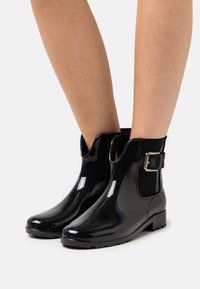 Bottes de pluie noires brillantes jusqu'à la cheville avec boucle sur le côté, portées sur des jambes nues devant un fond blanc uni.