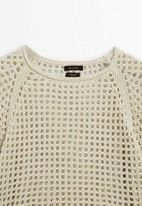 Hellbeige Baumwollpullover mit offenem Strickmuster, rundem Ausschnitt und Raglanärmel. Etikett am Hals sichtbar mit Marken- und Materialangaben.