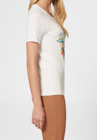 Camiseta blanca de manga corta con un diseño impreso colorido en el frente, combinada con pantalones ajustados de color marrón claro.