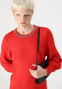 Pull en tricot rouge à manches longues, avec un col orné de bijoux. Le modèle tient un sac à main noir en texture lisse avec une bandoulière réglable.
