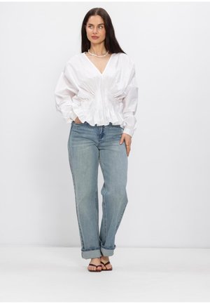 Woman wearing a white V-neck blouse with gathered waist, light blue cuffed jeans, black sandals, and a pearl necklace, standing with hands in pockets.