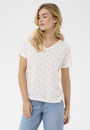 Blonde vrouw die een wit T-shirt met korte mouwen en een V-hals draagt, bedrukt met kleine zwarte polka dots, en lichtblauwe spijkerbroek, poserend met ontspannen handen.
